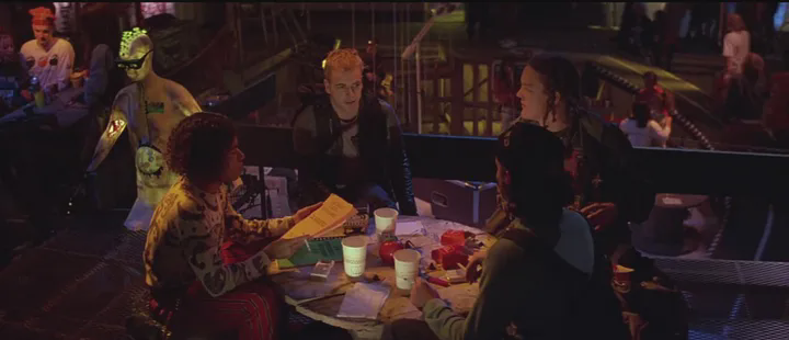 Group of youg man sitting at a table, covered in coloroful ligting. They are talking and holding and orange book.