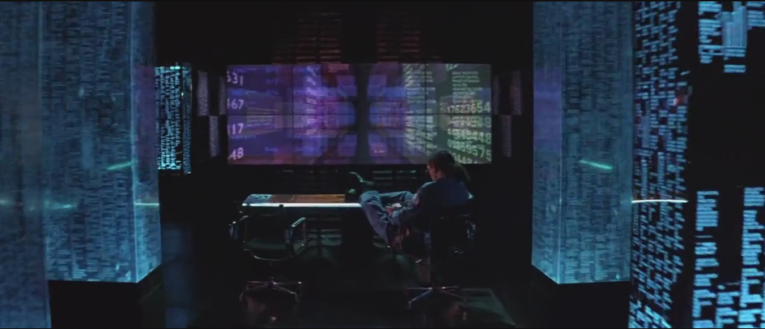 An white man is sitting in the center, back to us. In front of him is a huge screen with blue city-scape is in front of him.