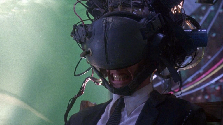 Keanu Reeves in metal VR goggles grimming his teeth. The helmet covers most of his face.