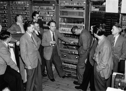 Black and white photo of a group of man standing next to a mechanical computer