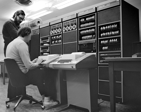 Photo of two white men. One is standing over and talking the other. The other is sitting in front of a teletype.